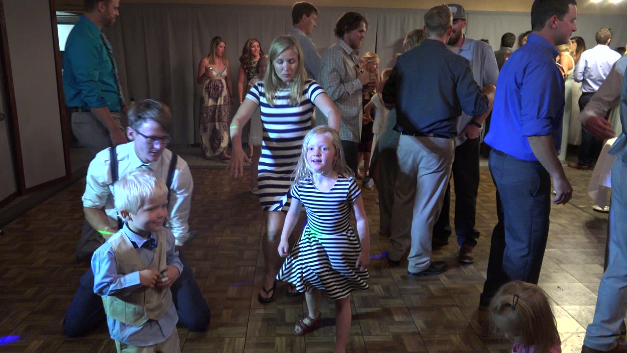 At 0:01 in Mesa, Colorado, a young boy, a kneeling adult, a girl in a striped dress, and several standing adults fill the indoor dance floor under scattered green and blue lights.