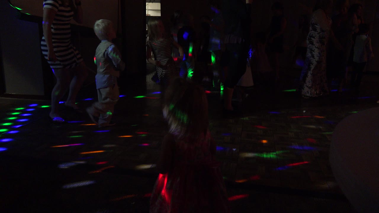 At the start of the Mesa, Colorado clip, children and adults stand and dance in a dim indoor room with red, green, and blue disco spots on the floor.