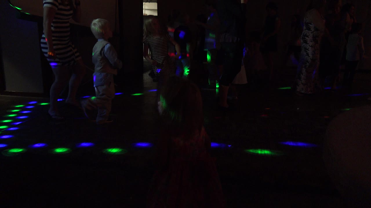 A young child moves across the Mesa, Colorado dance floor while adults and other children remain in the darker background under green and blue light spots.