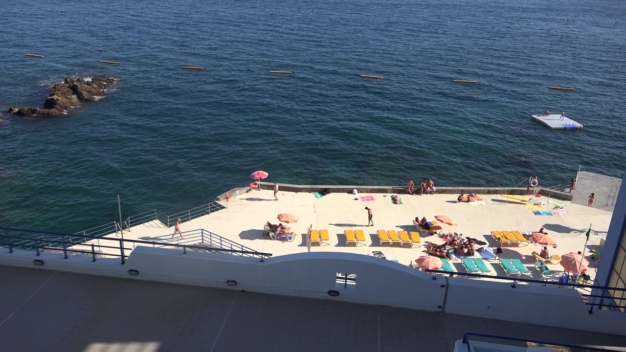 High-angle Madeira Portugal frame showing a white sun deck with loungers, blue railings, rocky outcrops, open ocean, and distant people on the terrace.