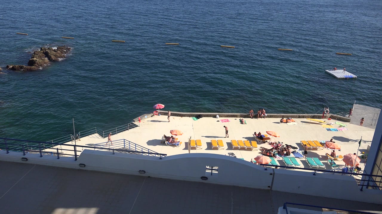 Madeira Portugal shoreline view with the sun deck stretched across the lower frame, loungers in bright sun, distant people near umbrellas, and rocks in the water.