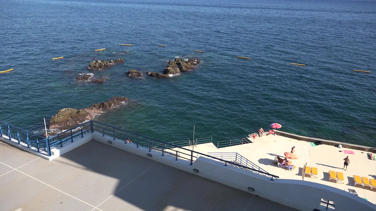 Madeira coastal view with the concrete terrace across the lower frame, blue railings at the edge, swimmers or beachgoers appearing very small, and dark rocks surrounded by calm water.