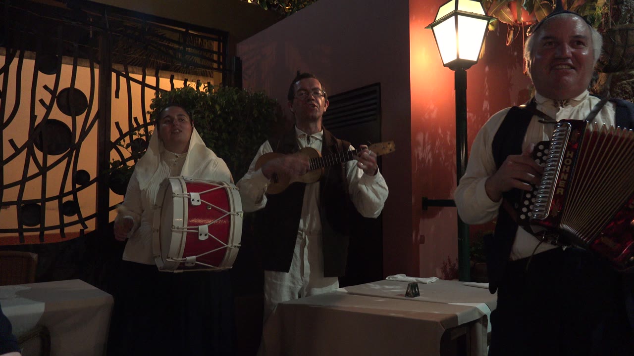 Three adult musicians perform in Madeira, Portugal, with a red accordion player near the camera and a drummer and guitarist behind him under warm indoor lighting.