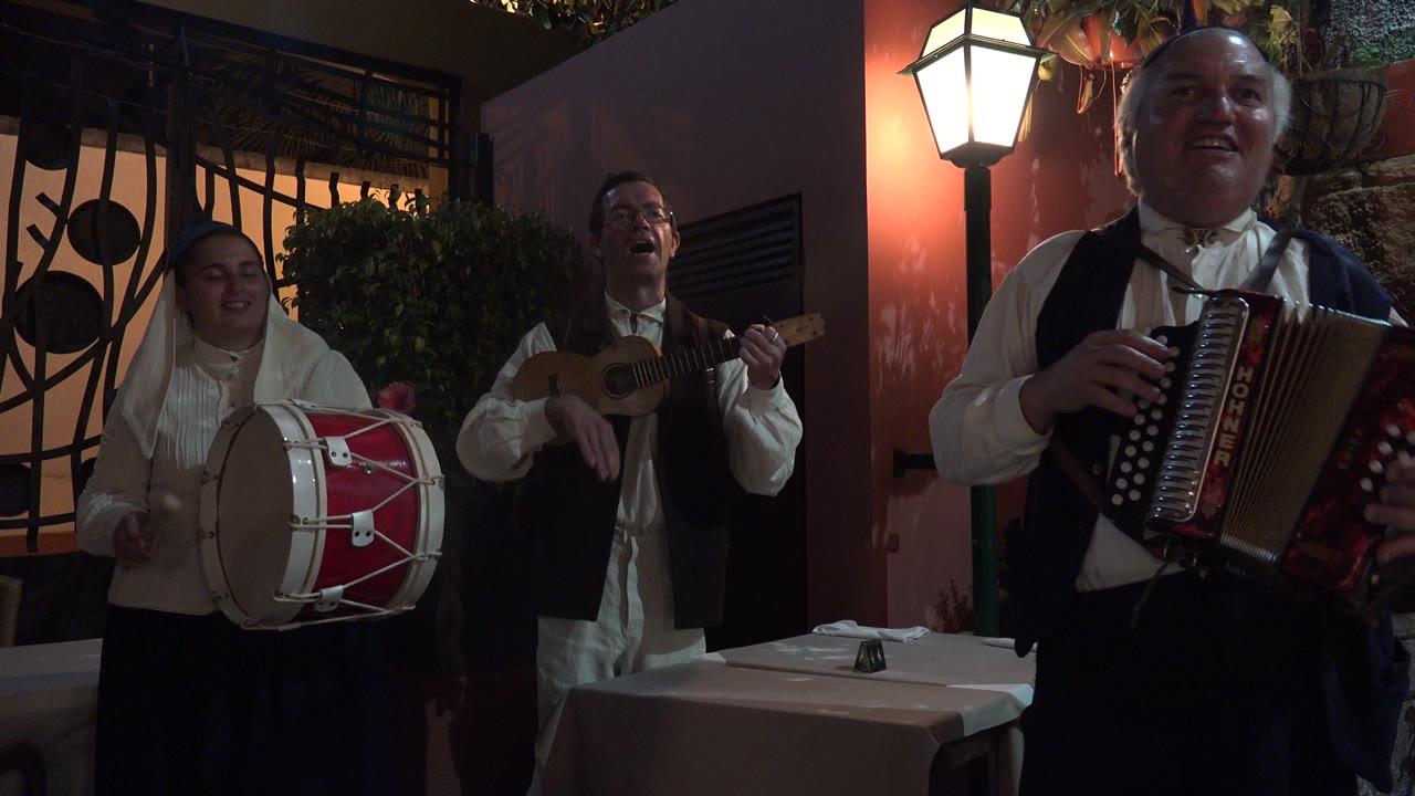 Three musicians in white shirts play drum, small guitar, and accordion under a wall light.