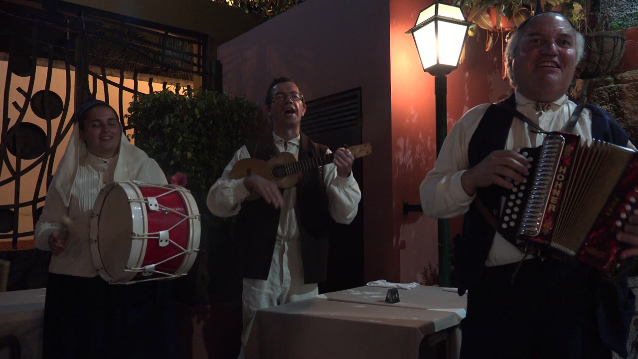 The accordion player fills the right foreground in Madeira, Portugal, while the drummer and guitarist remain visible behind him near a lit wall and dark ironwork.
