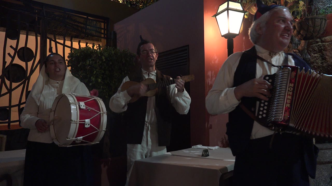 Three musicians perform at night with a drum, small string instrument, and accordion.