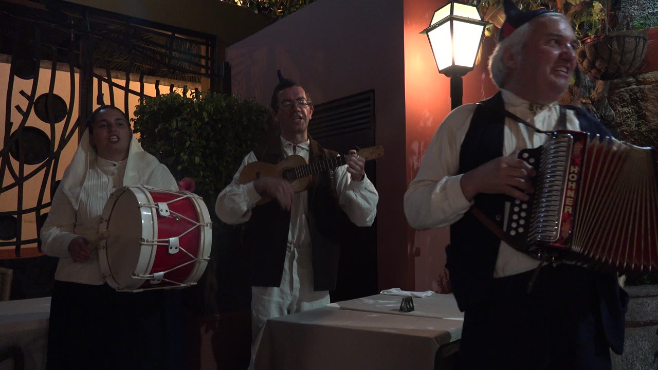 In Madeira, Portugal, the accordion player dominates the right foreground while the drummer and string player remain behind him beside a lamp-lit pink wall and barred window.