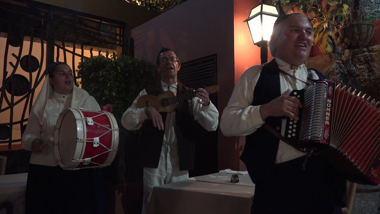 Three adult musicians performing outdoors at night with drum, small guitar, and accordion.