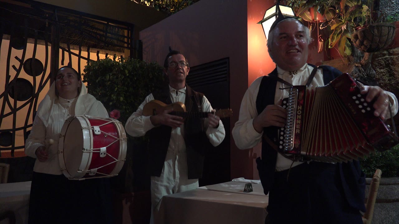 Three folk musicians performing with drum, guitar-like instrument, and accordion.