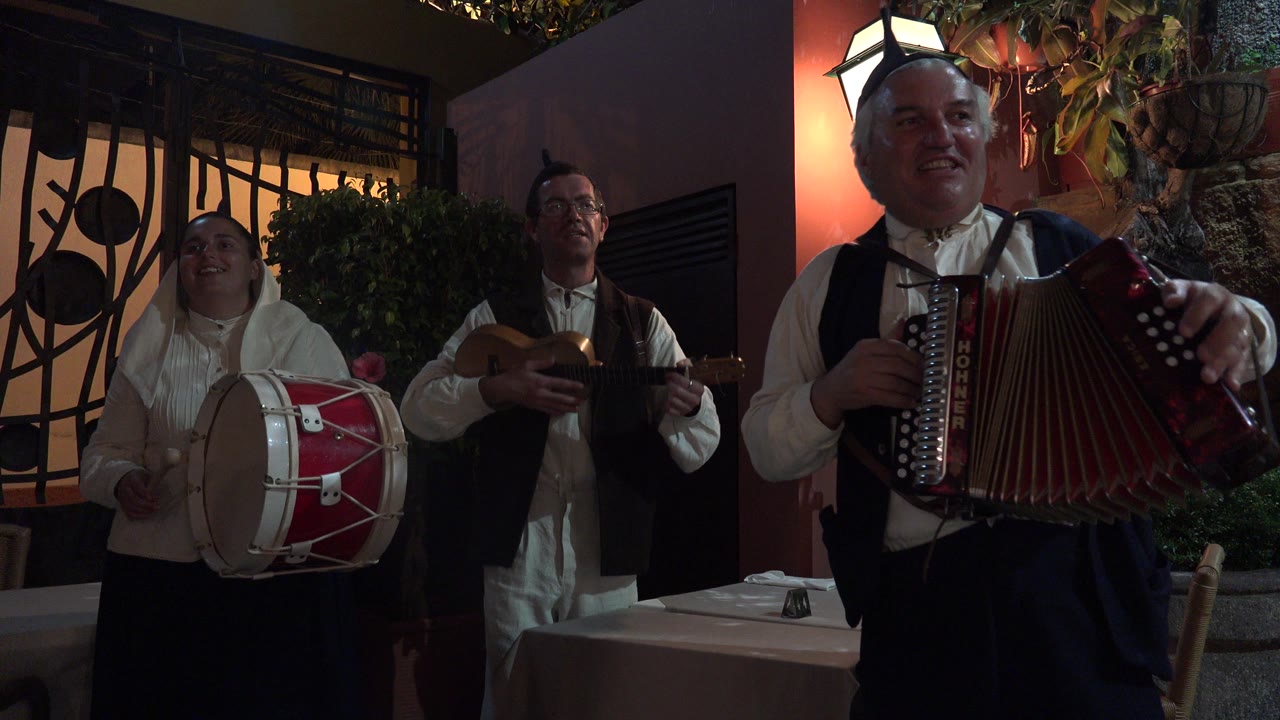 A smiling accordion player stands close to the camera in Madeira, Portugal, with the drummer and guitar player behind him near a barred window and a lit wall lamp.