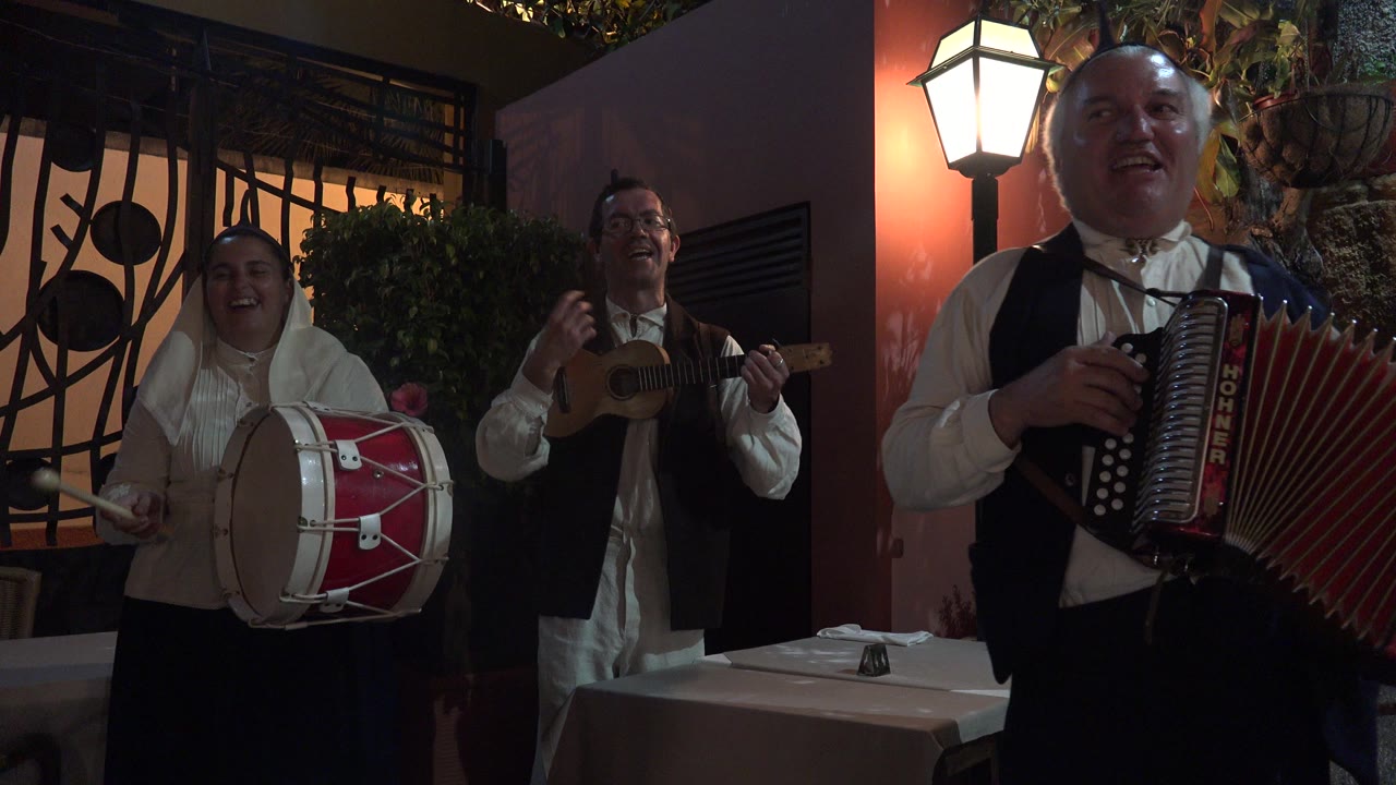 The accordion player fills the right foreground while the drummer and guitarist remain visible behind him in the warm-lit Madeira restaurant interior.