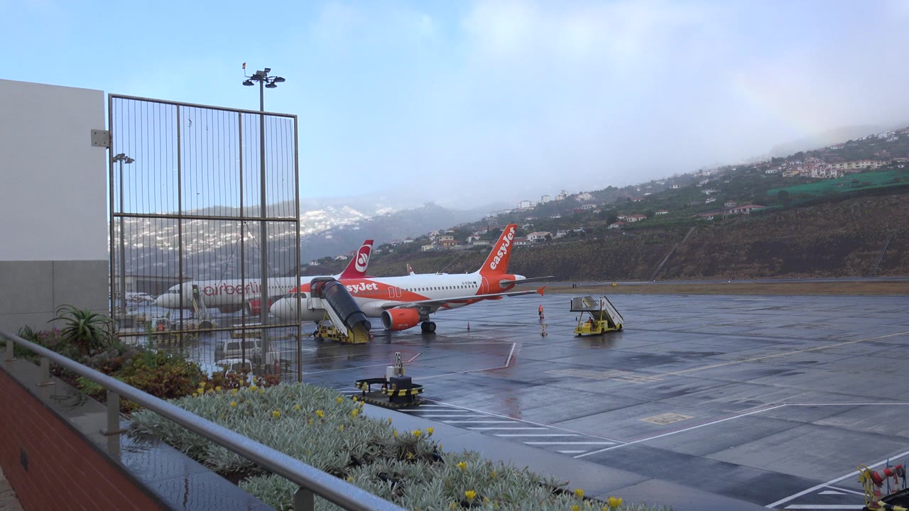 Opening frame of a Madeira, Portugal airport apron with an easyJet aircraft beside service stairs, wet pavement, railing and plants in the foreground, hillside buildings in back, and no visible people.