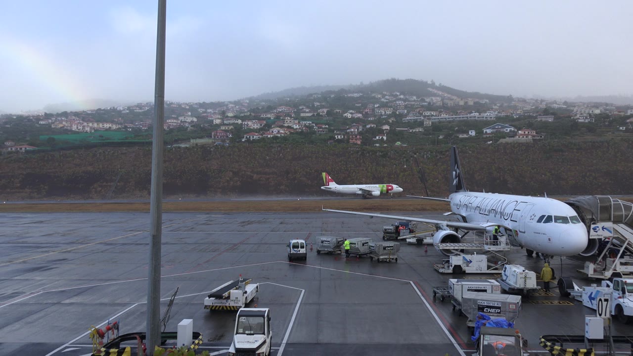 A Madeira, Portugal airport apron shows a white commercial aircraft parked beside stairs and ground equipment, with wet pavement, low cloud, and hillside homes behind it.