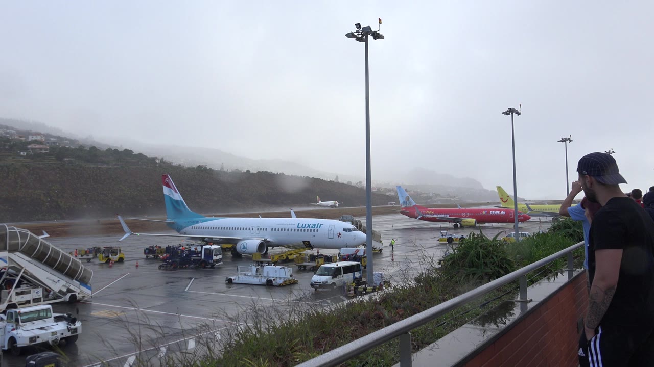 Airport apron with aircraft, service vehicles, railing, and a man on a viewing terrace.