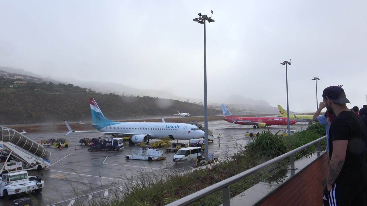 Madiera Portugal airport view with the Luxair aircraft centered lower in frame, red aircraft behind it, wet pavement, service equipment, tall lamps, and mist over the hills.