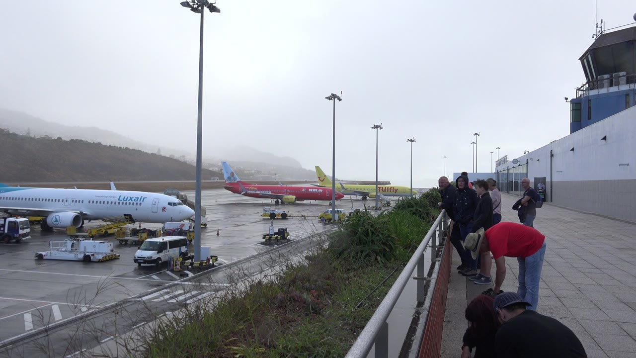 Travelers beside an airport terrace railing overlooking parked passenger jets.