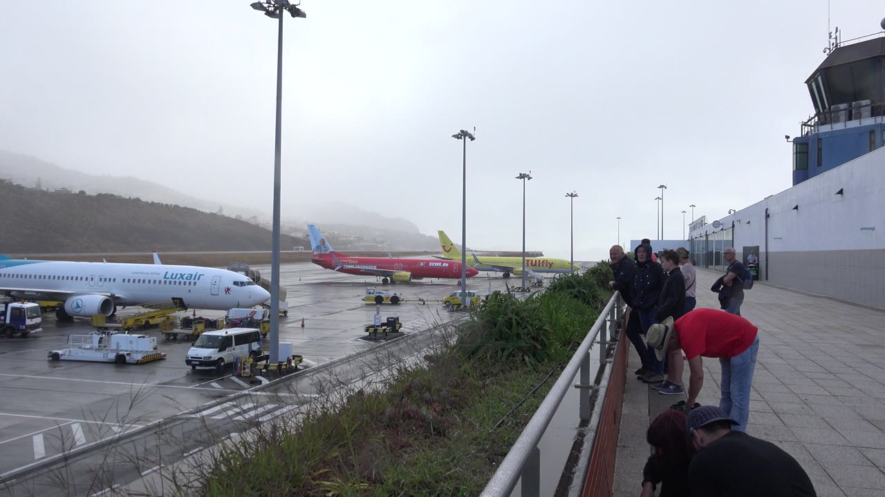 A similar terrace view in Madeira, Portugal shows passengers beside the railing as ground equipment sits around a Luxair jet and other aircraft on the wet apron.