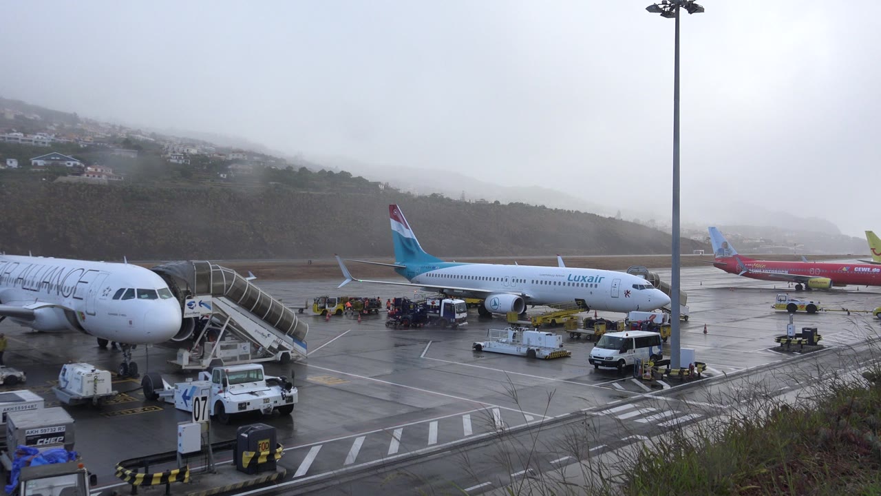 Madeira Airport ramp view with a white jet at left, a Luxair plane in the midground, baggage carts, service vehicles, and gray mist over the hillside.