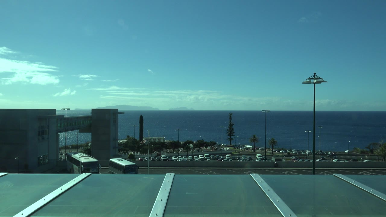 Opening frame of Madeira, Portugal with a cool blue ocean horizon, distant waterfront road, low buildings, parked cars, and roof panels in the foreground, with no visible people.
