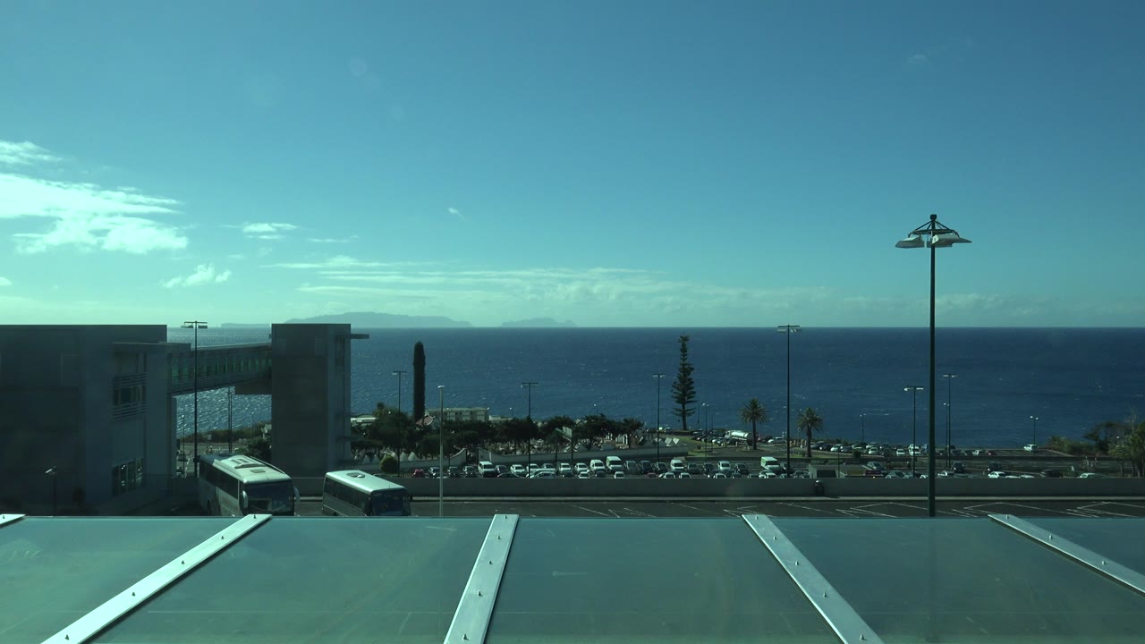 Opening frame of a Madeira, Portugal sea view from an elevated building, with roof panels in front, parked cars and buses below, ocean beyond, and no visible people.