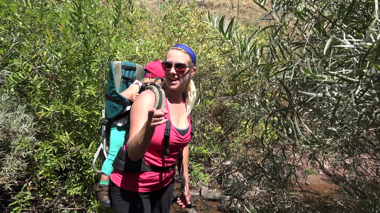 Smiling woman carrying a child on her back beside green shrubs.