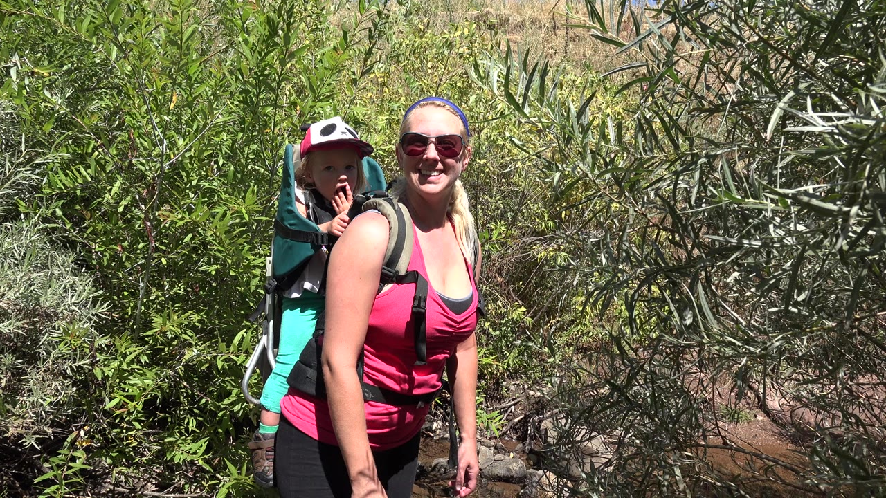 A woman in sunglasses carries a young child in a backpack carrier while standing near dense green shrubs in Golden, Colorado.