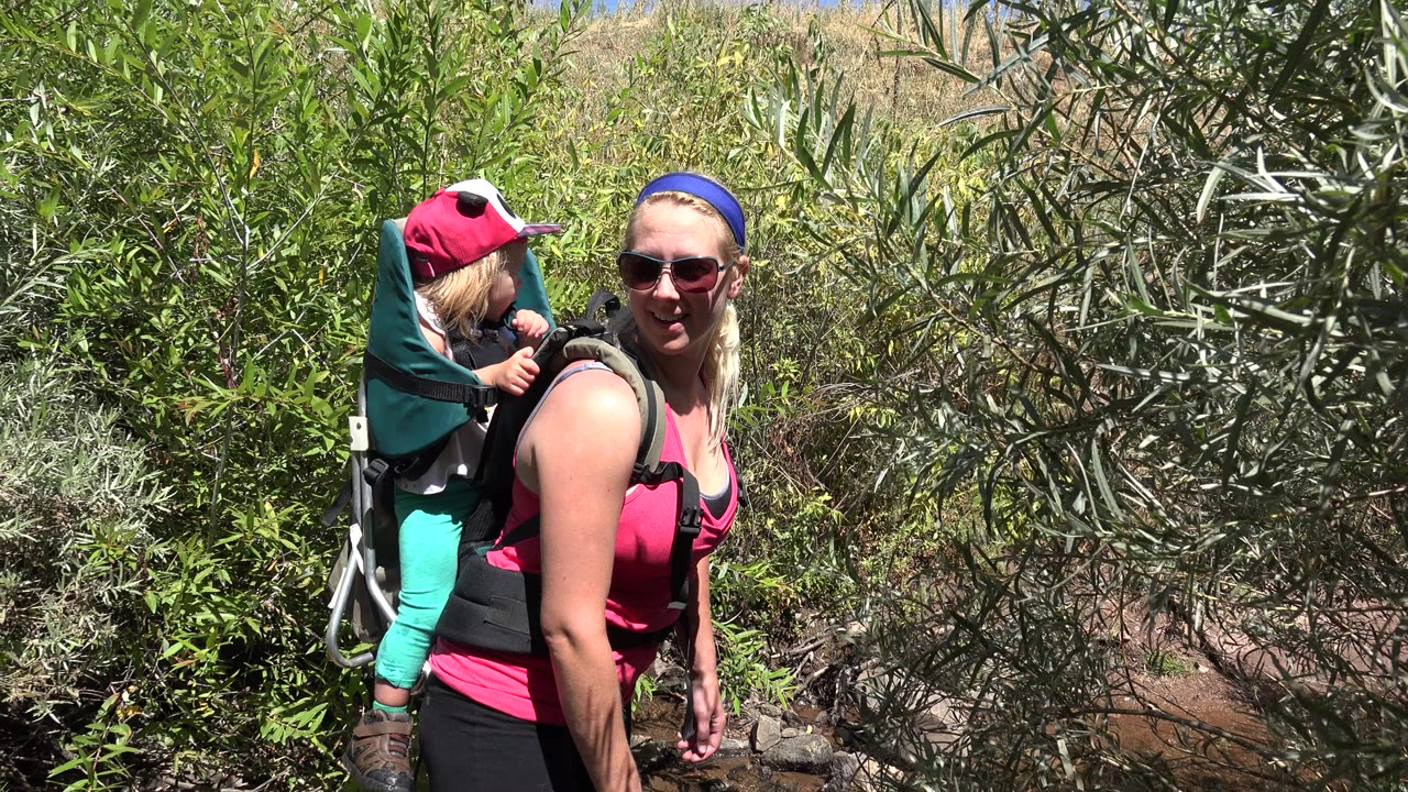In Golden, Colorado, a blonde woman in sunglasses stands near green brush with a small child seated in a backpack carrier behind her.