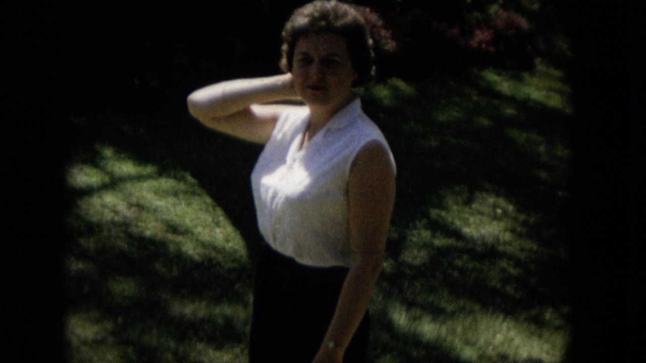 Adult woman in a white sleeveless shirt and dark shorts stands on dappled grass in Keenesburg, Colorado, with her arm raised near her neck.
