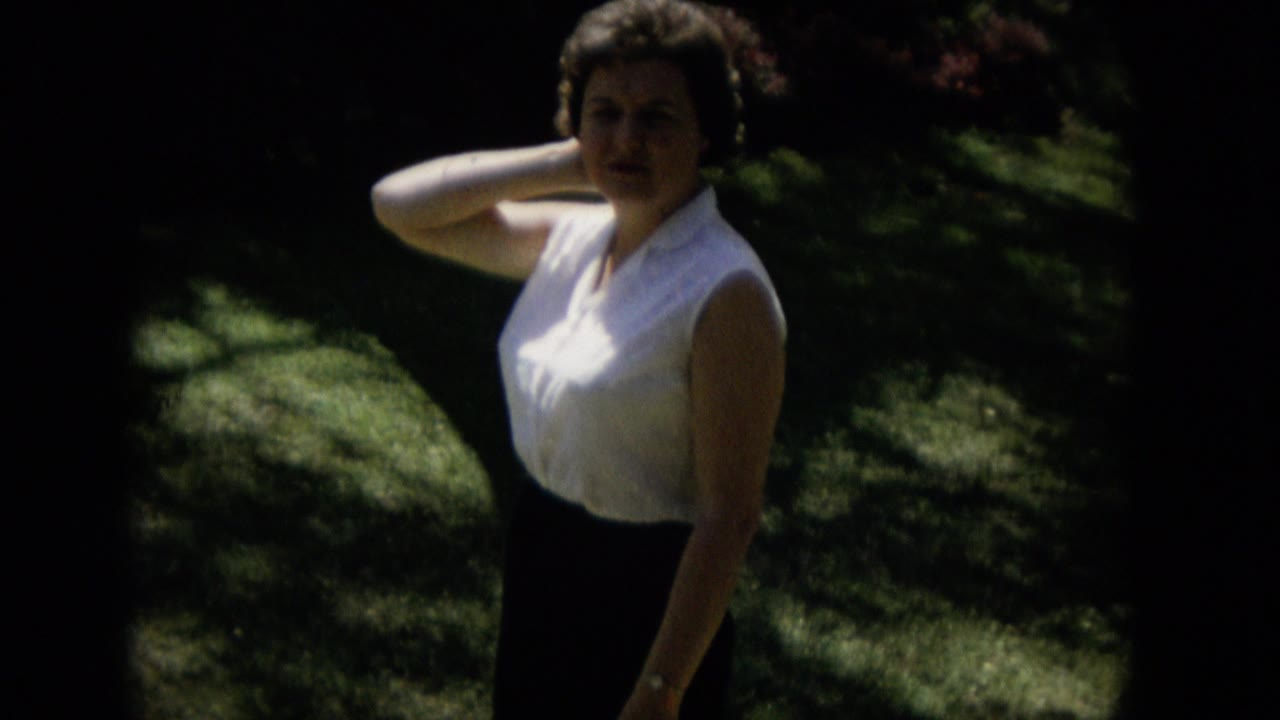 The woman remains in a white shirt and dark shorts on shaded grass in Keenesburg, Colorado, framed from head to knees with her arm still near her neck.