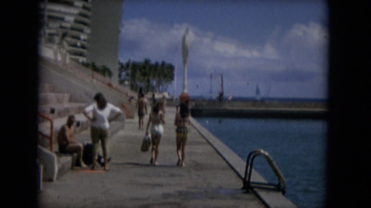People in swimsuits walk along a concrete seawall beside blue water.