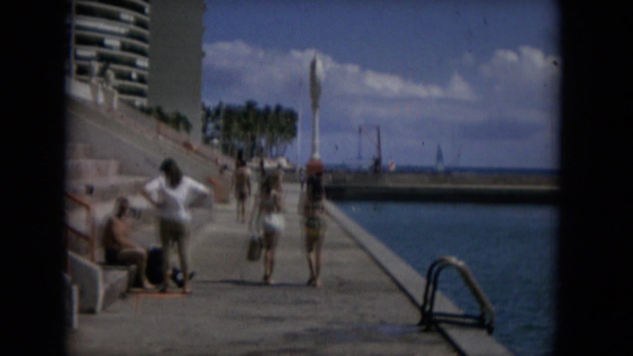 A small group of swimsuit pedestrians moves farther along the Keenesburg, Colorado seawall, with seated people at left, water at right, and palm trees beyond the walkway.