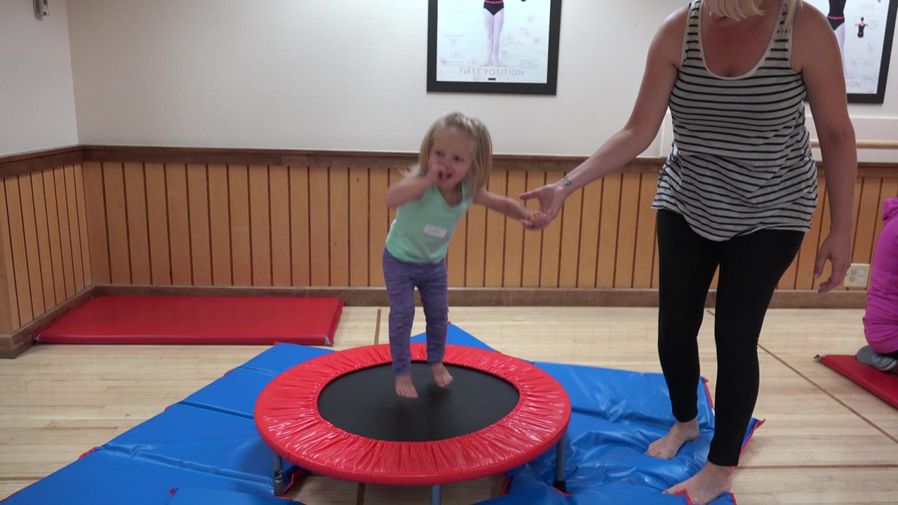 At 0:00 in Westminster, Colorado, a young child leans forward on a red-and-black mini trampoline while holding an adult woman's hand beside blue floor mats.