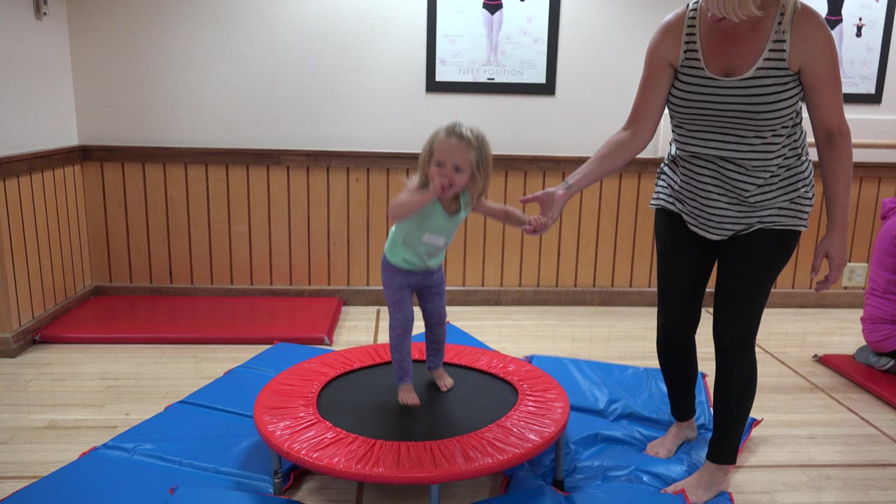 At 0:01 in Westminster, Colorado, the child is mid-bounce on the mini trampoline, one hand raised near her face and the other held by the adult woman.