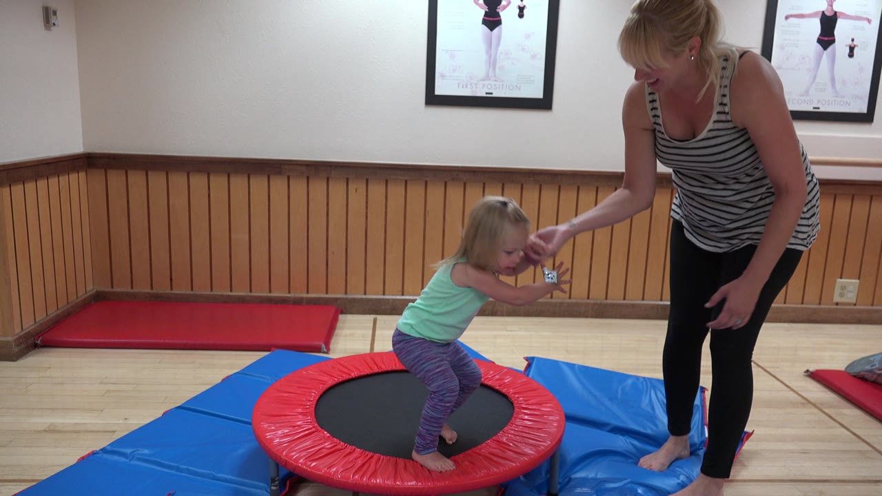 Young child on a red trampoline holds an adult woman's hands indoors.