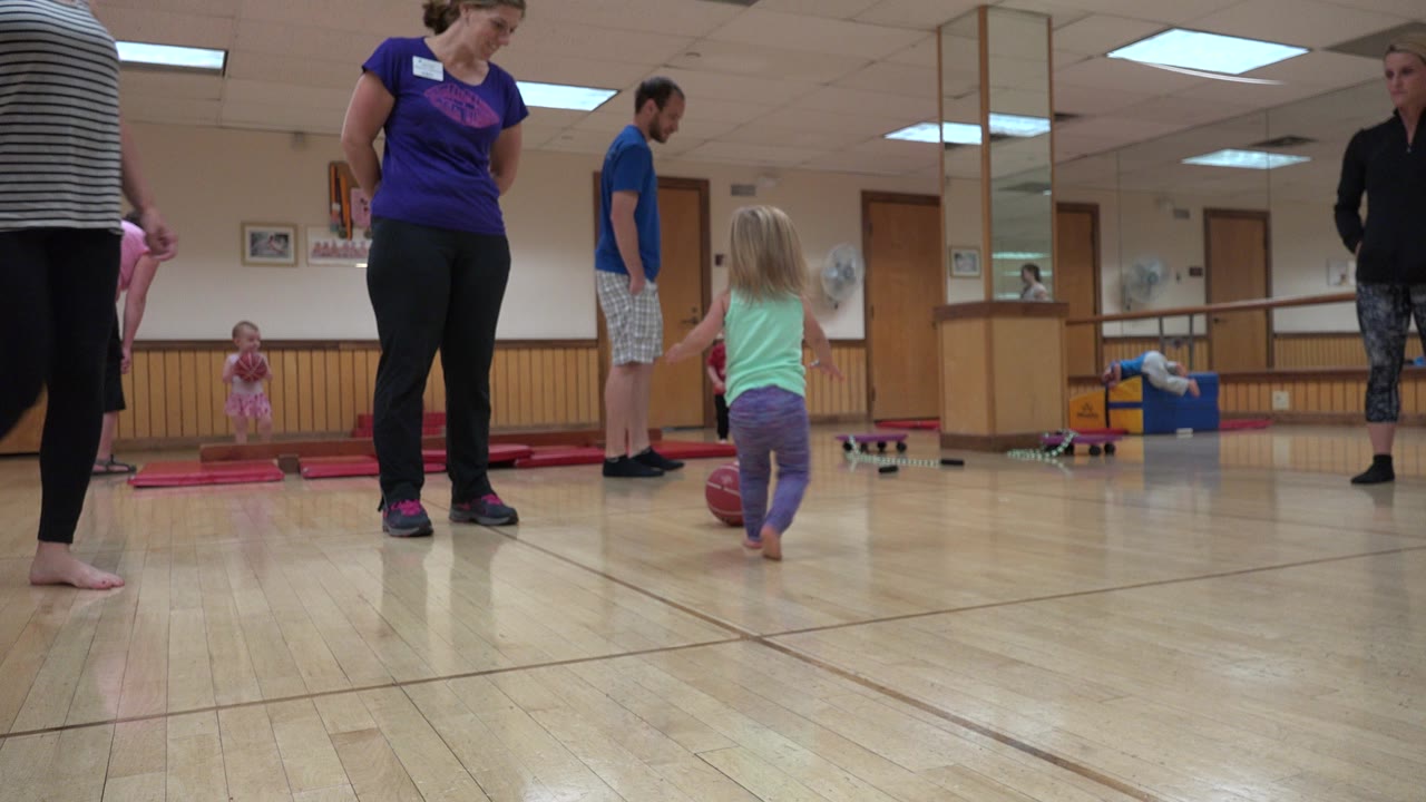 Girl approaches a red ball in an indoor play room with adults nearby.