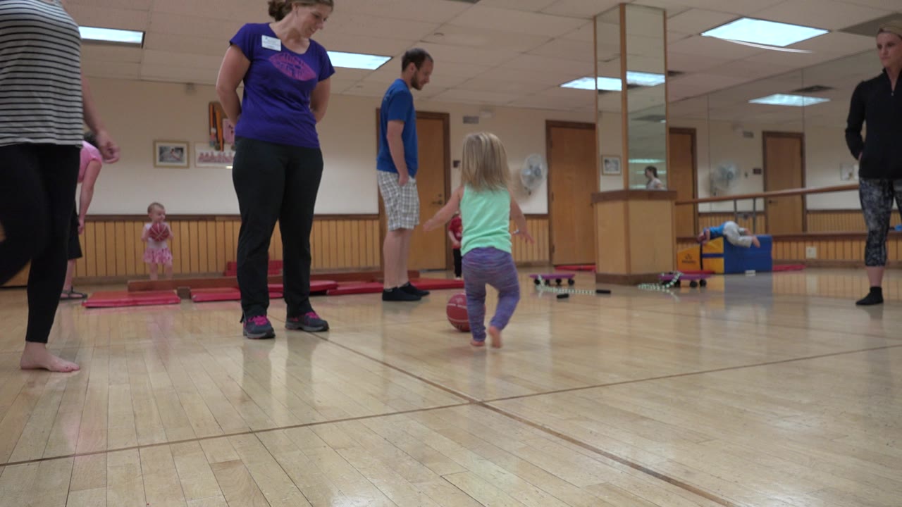 The girl continues toward the red ball in the Westminster indoor play space, with an adult in a purple shirt, a man in blue, and children visible near mats and toys.