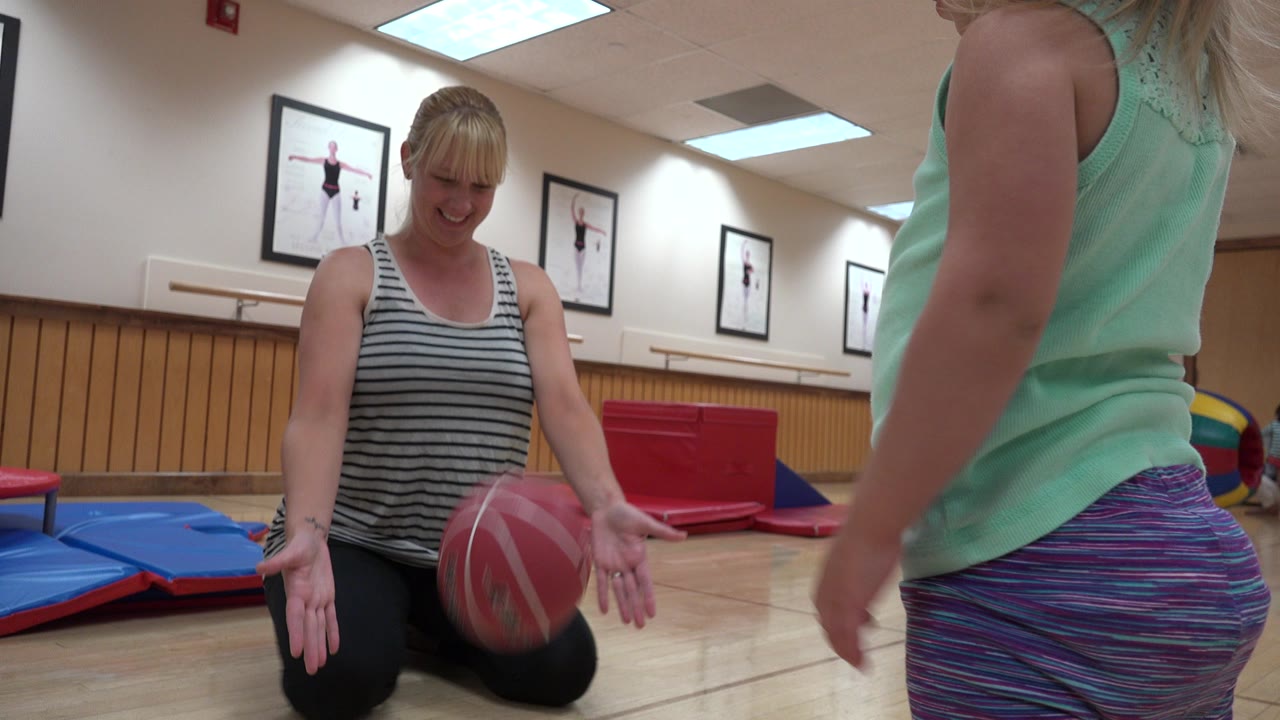 Woman kneeling with a pink ball near a standing child in an indoor gym room.