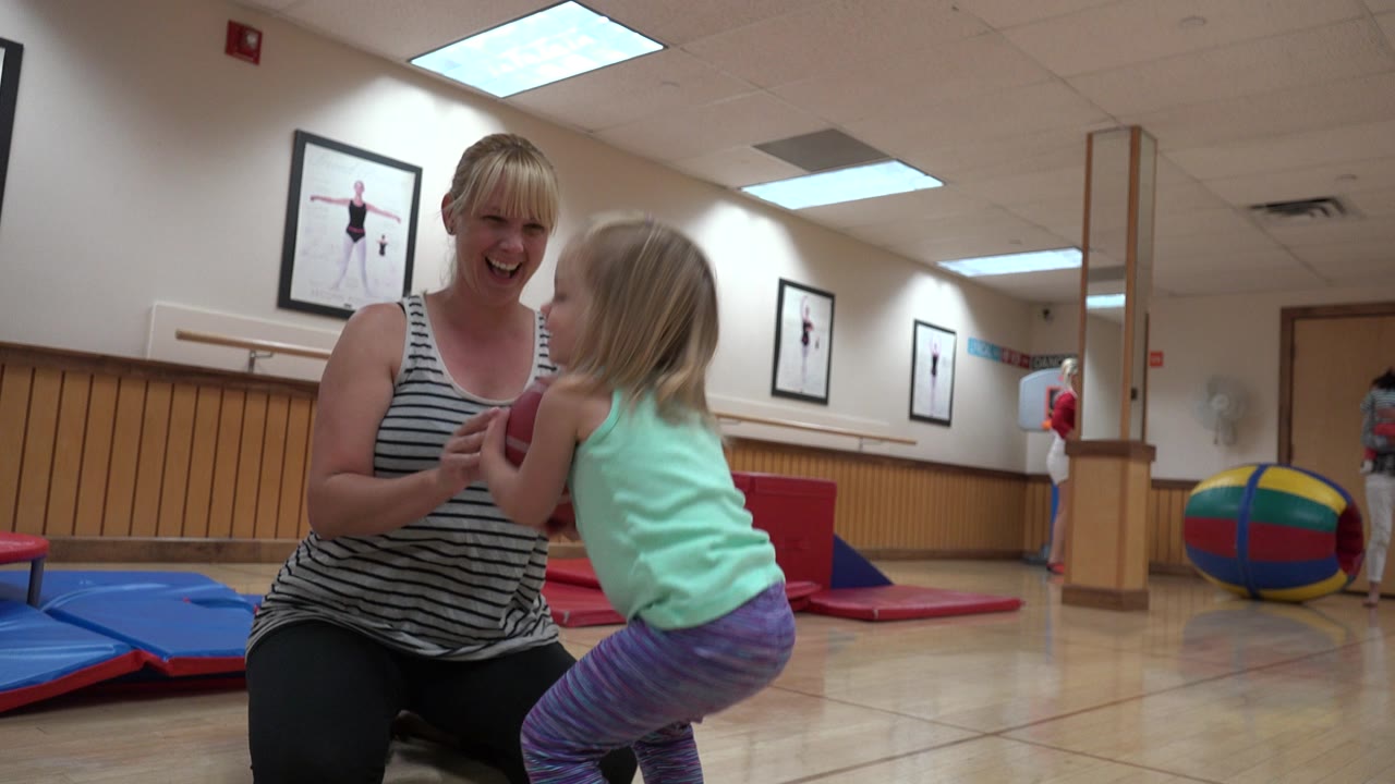 In Westminster, Colorado, a kneeling mother smiles near a blonde toddler holding a red ball in a gym with mats and framed posters behind them.