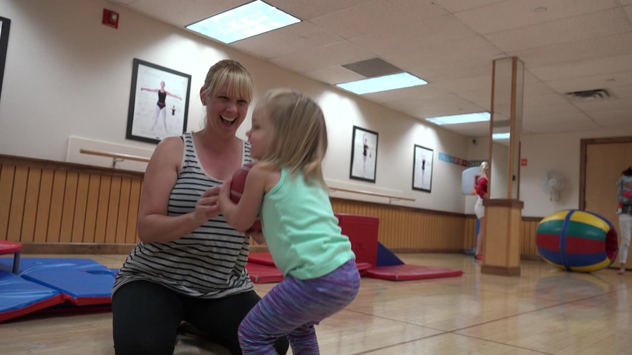 In Westminster, Colorado, the toddler faces the kneeling mother with the red ball near her chest, while blue mats and a large multicolor ball sit in the gym background.
