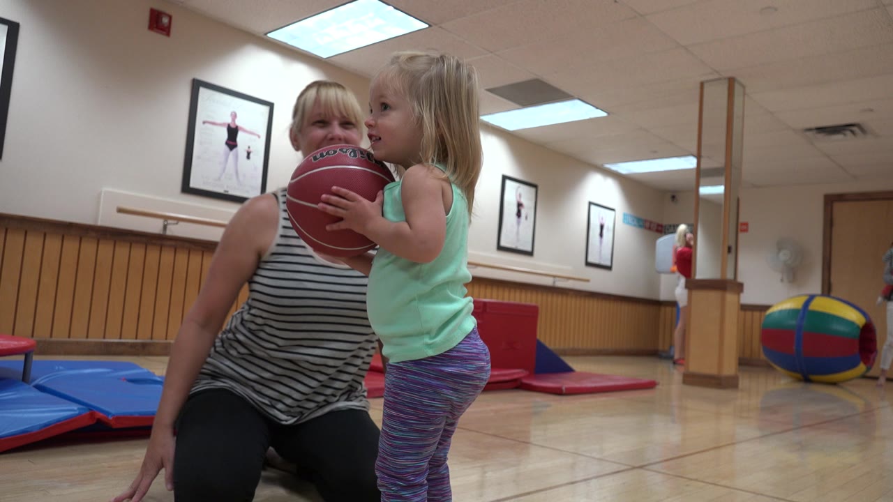 In Westminster, Colorado, the toddler stands close to the kneeling mother with the red ball lifted near her shoulder, with gym mats and wall posters visible behind them.