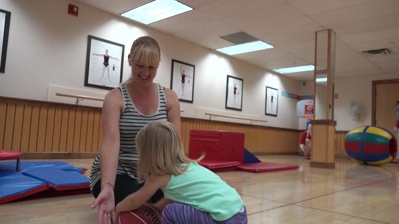 Young girl reaches toward a brown ball beside a kneeling woman in a gym.