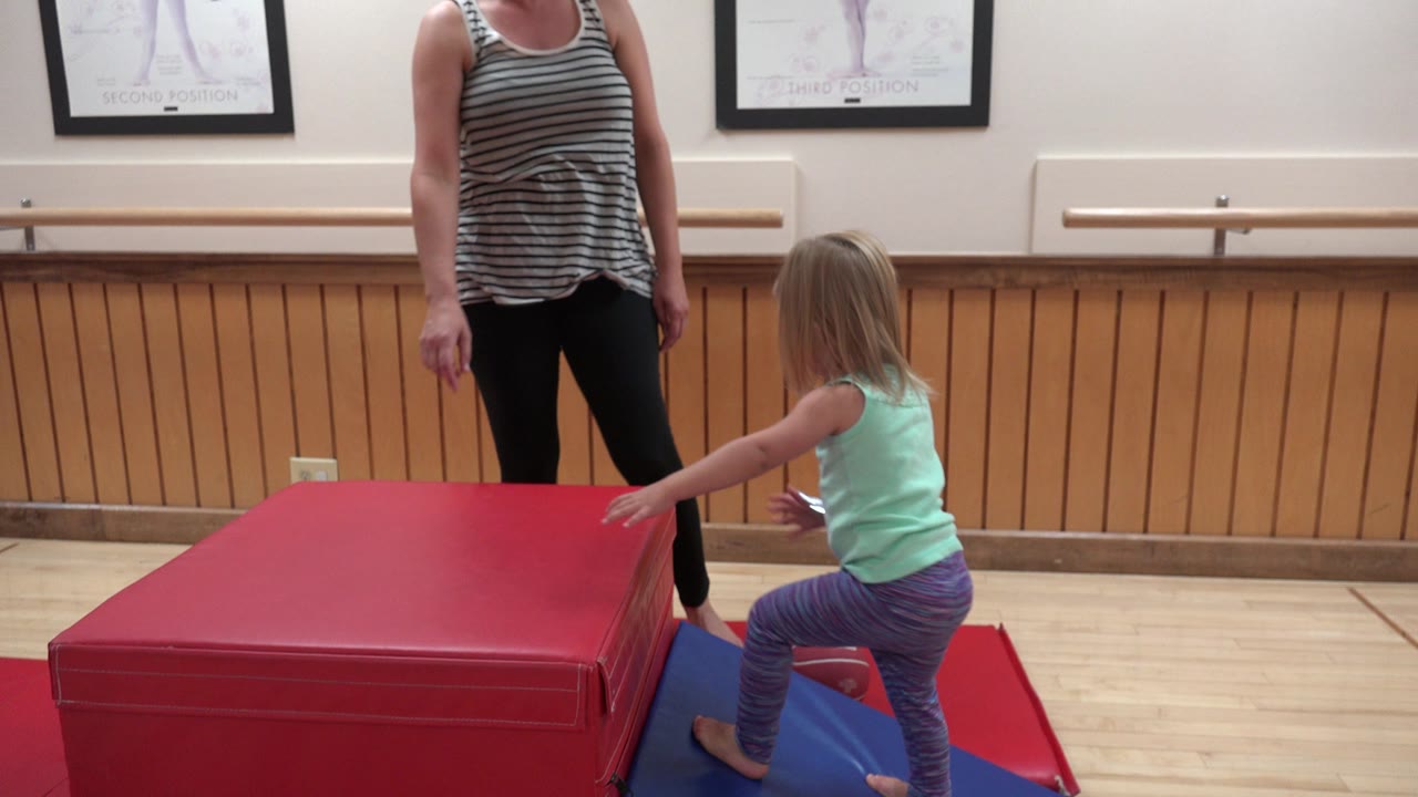 In Westminster, Colorado, the girl leans forward with one foot near the blue mat and one hand reaching toward the red padded block while her mother stands in the background.