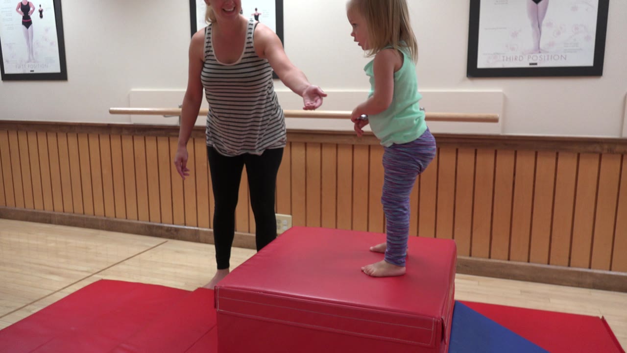 The child faces the woman while standing on the red block in the Westminster gym, with bright indoor light on the mats and framed exercise posters on the wall.