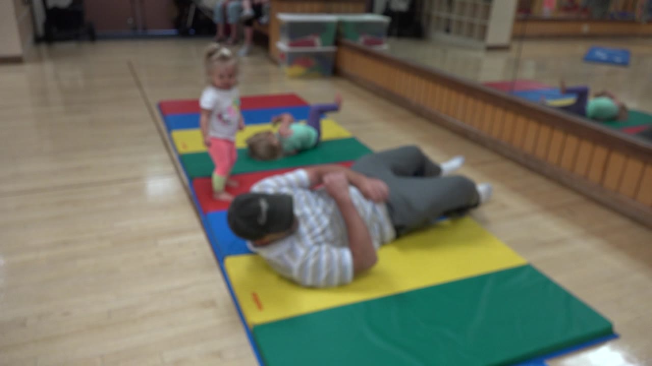 The father stays curled on the yellow section of the mat while the toddler girl stands upright beside the red and blue panels in the Westminster indoor play space.