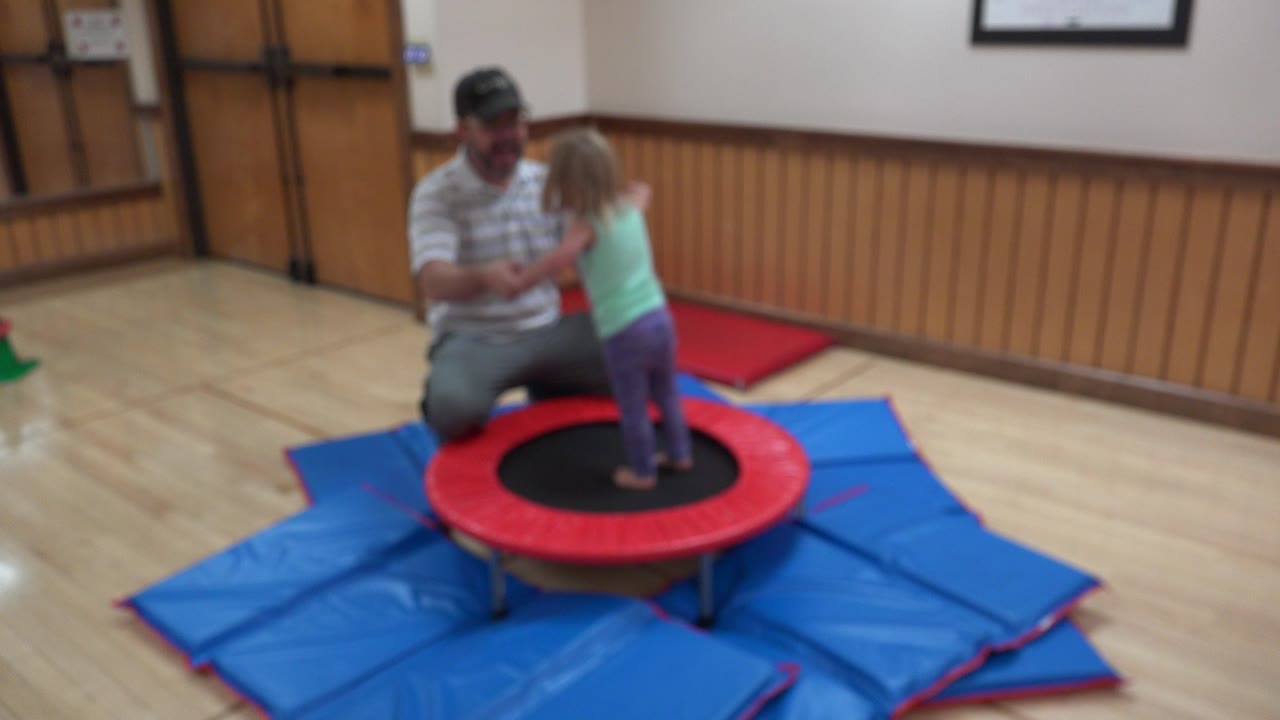In Westminster, Colorado, a man kneels beside a young girl standing on a red-and-black indoor trampoline set on blue mats.