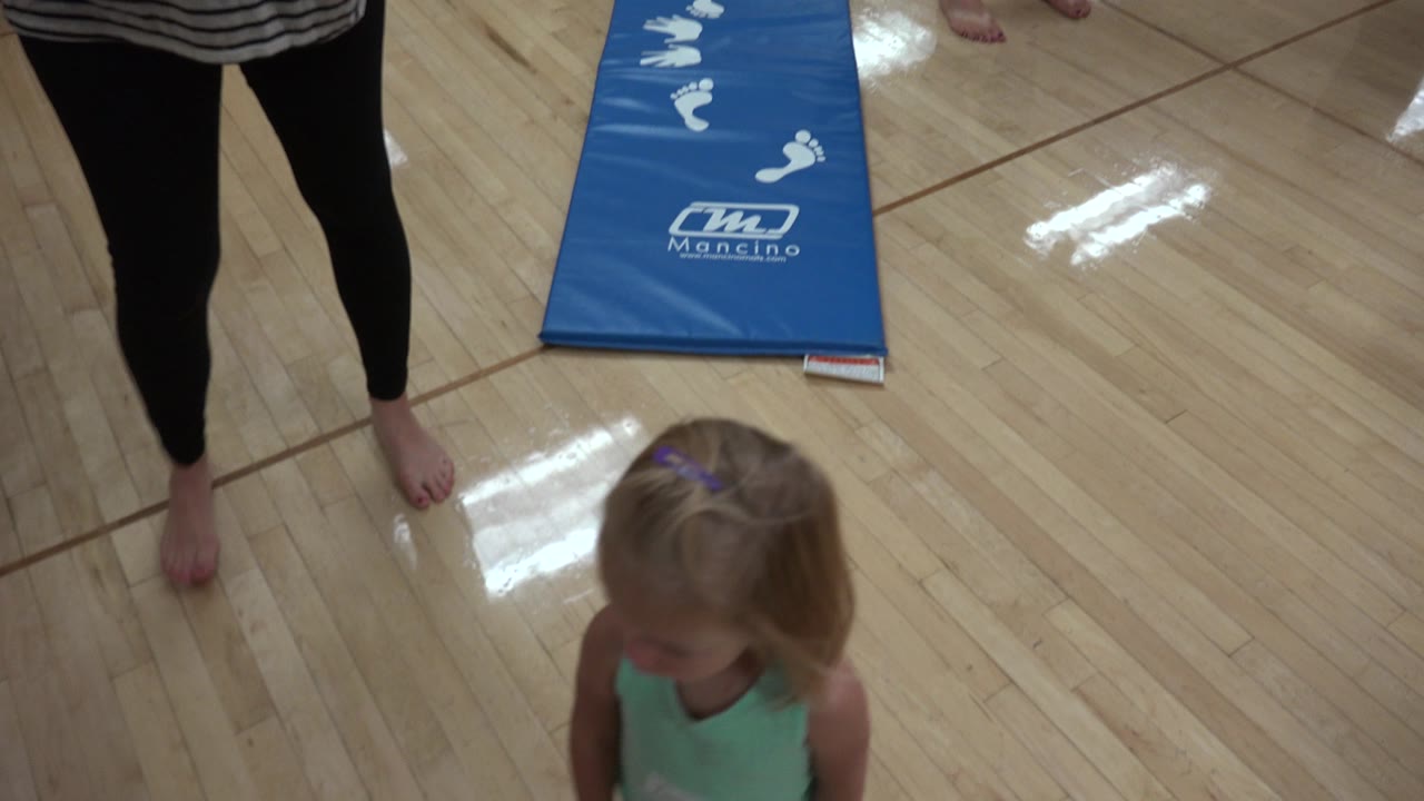 In Westminster, Colorado, a young girl stands near an adult's bare feet on a shiny indoor floor with a blue exercise mat behind her.