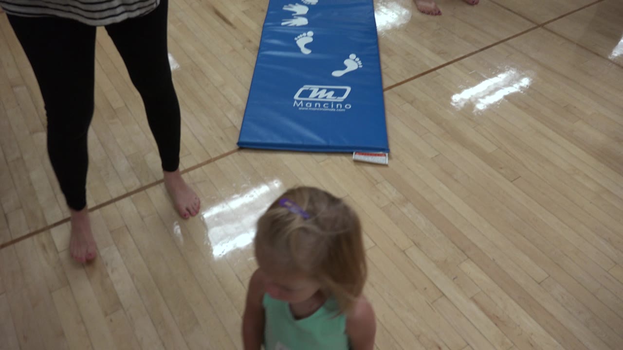 The young girl remains near the adult's legs on the glossy floor, with the blue mat and its white markings visible farther back in the Westminster indoor space.