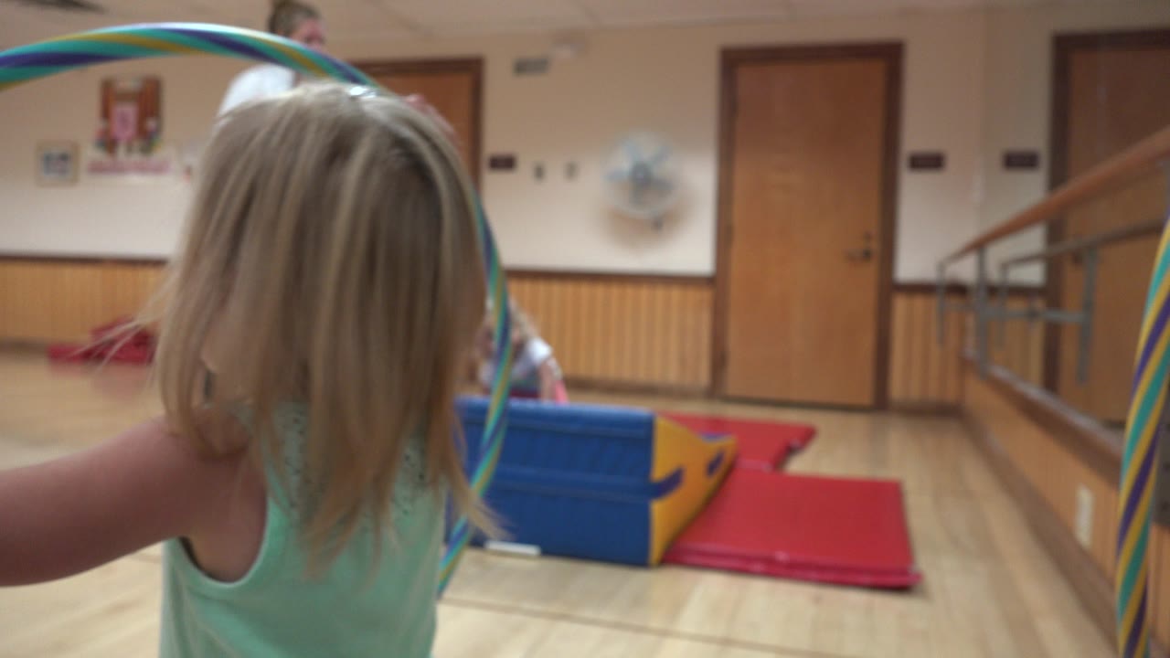 A blonde child in a mint tank top stands near the camera in Westminster, Colorado, holding striped ribbons in a mirrored indoor gym room with mats and a foam block ahead.