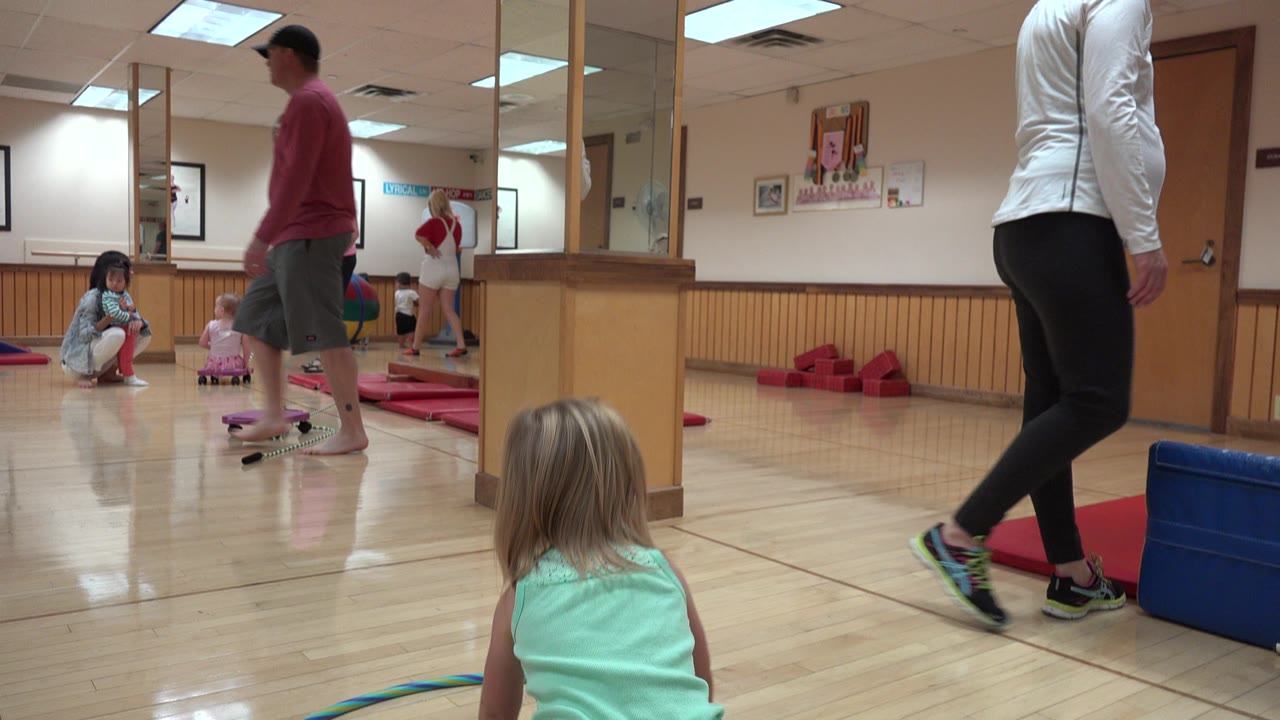 Young girl near a hula hoop on a gym floor with adults and children in the background.
