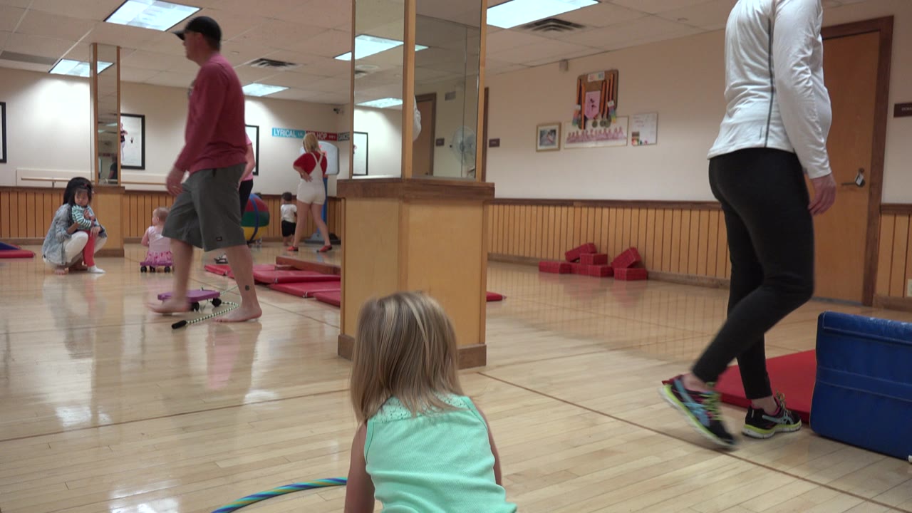 The young girl is seen from behind facing the hula hoop on the gym floor, with an adult in a white top close to the camera and other children and adults farther back in Westminster, Colorado.